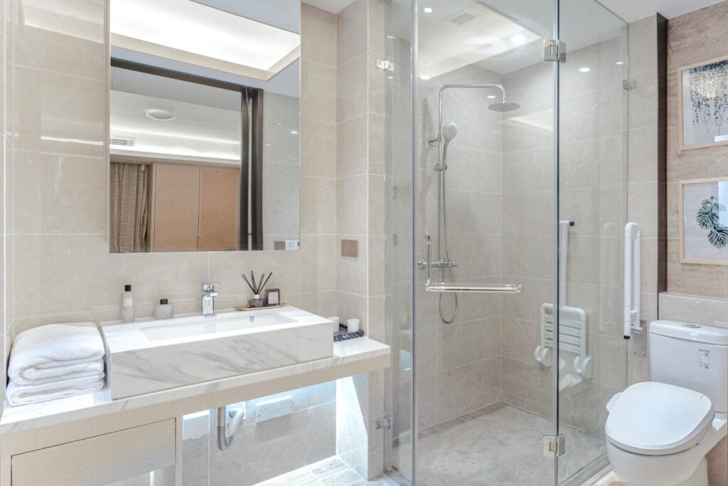Toilet Repair & Installation 4 Modern bathroom with a glass-enclosed shower, marble sink, toilet, and shelves with toiletries. Towels are stacked beside the sink, and framed art decorates the beige-tiled walls. Recently completed plumbing ensures everything runs smoothly in this elegant space.