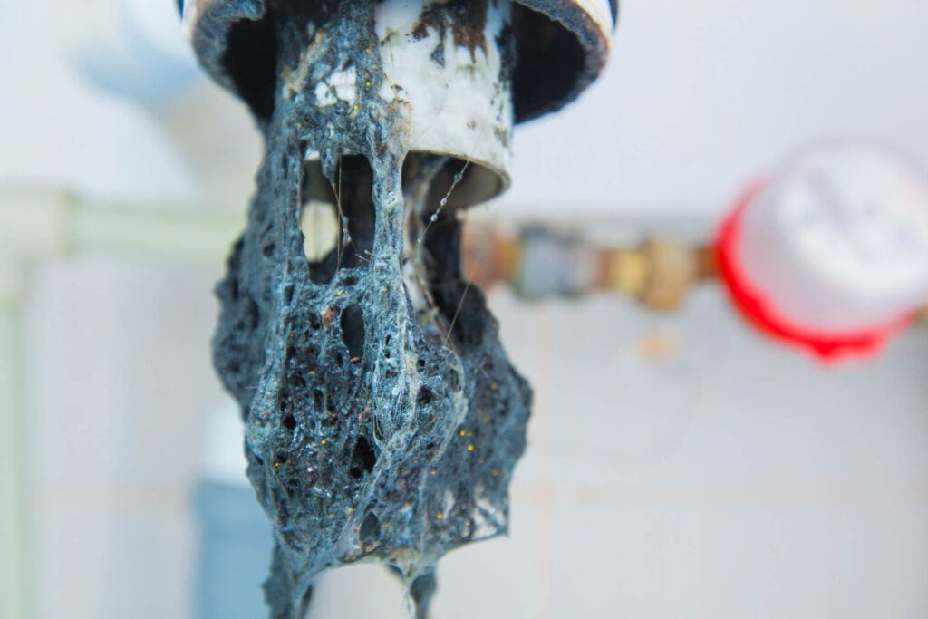 Toilet Repair & Installation 7 Close-up of a clogged pipe with dark, tangled debris hanging from the opening, reminiscent of a common toilet repair issue, set against a blurred background with faint red and white objects.