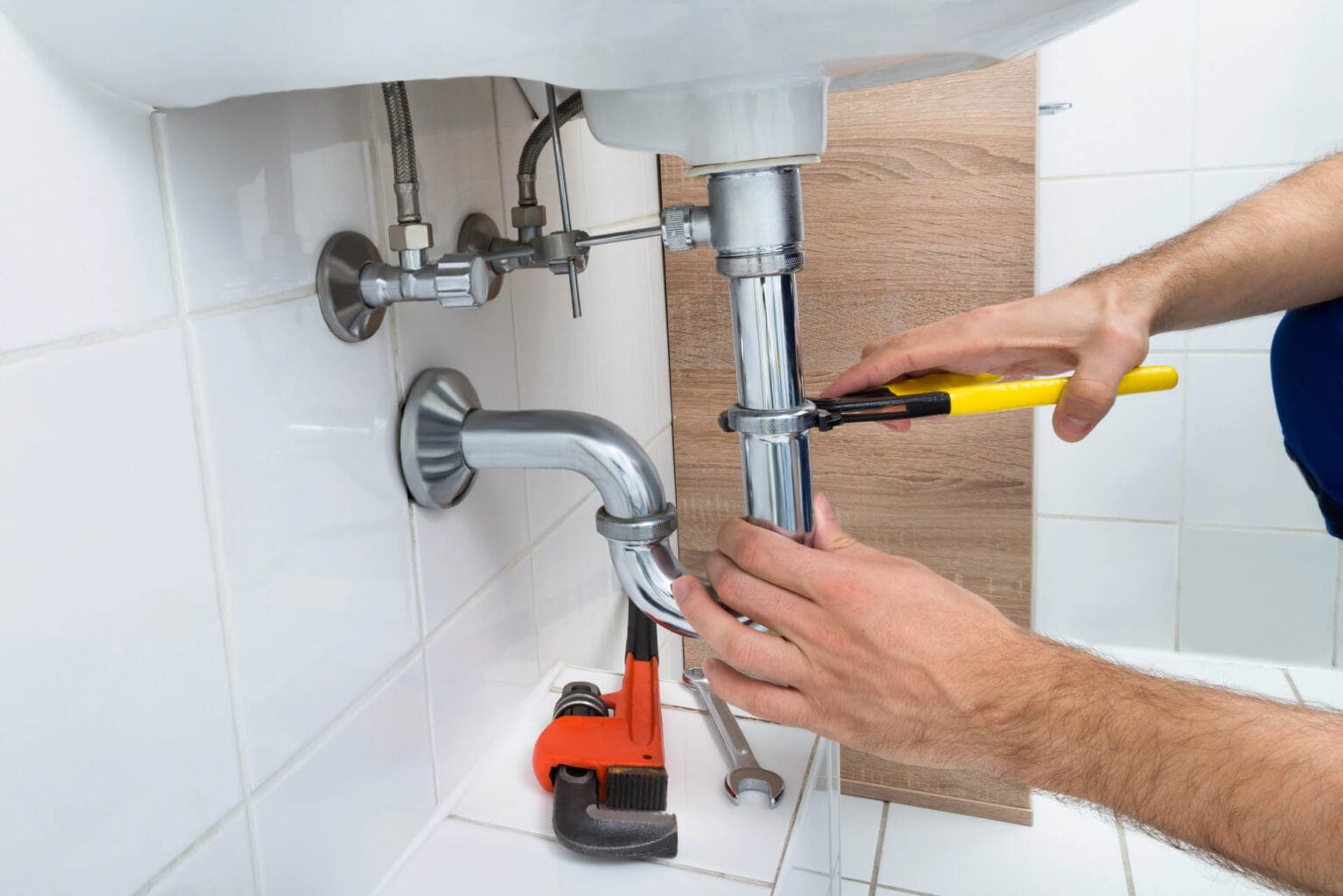 Toilet Repair & Installation 2 A person is skillfully handling plumbing work under a bathroom basin, using a yellow wrench to fix the sink pipe. Meanwhile, a red pipe wrench sits on the tiled floor, ready for any additional toilet repair tasks.
