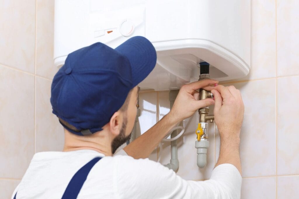 7 Signs It’s Time to Replace Your Water Heater 1 A person in a cap and overalls is carefully assessing when to replace water heater as they adjust the wall-mounted unit in a tiled room.