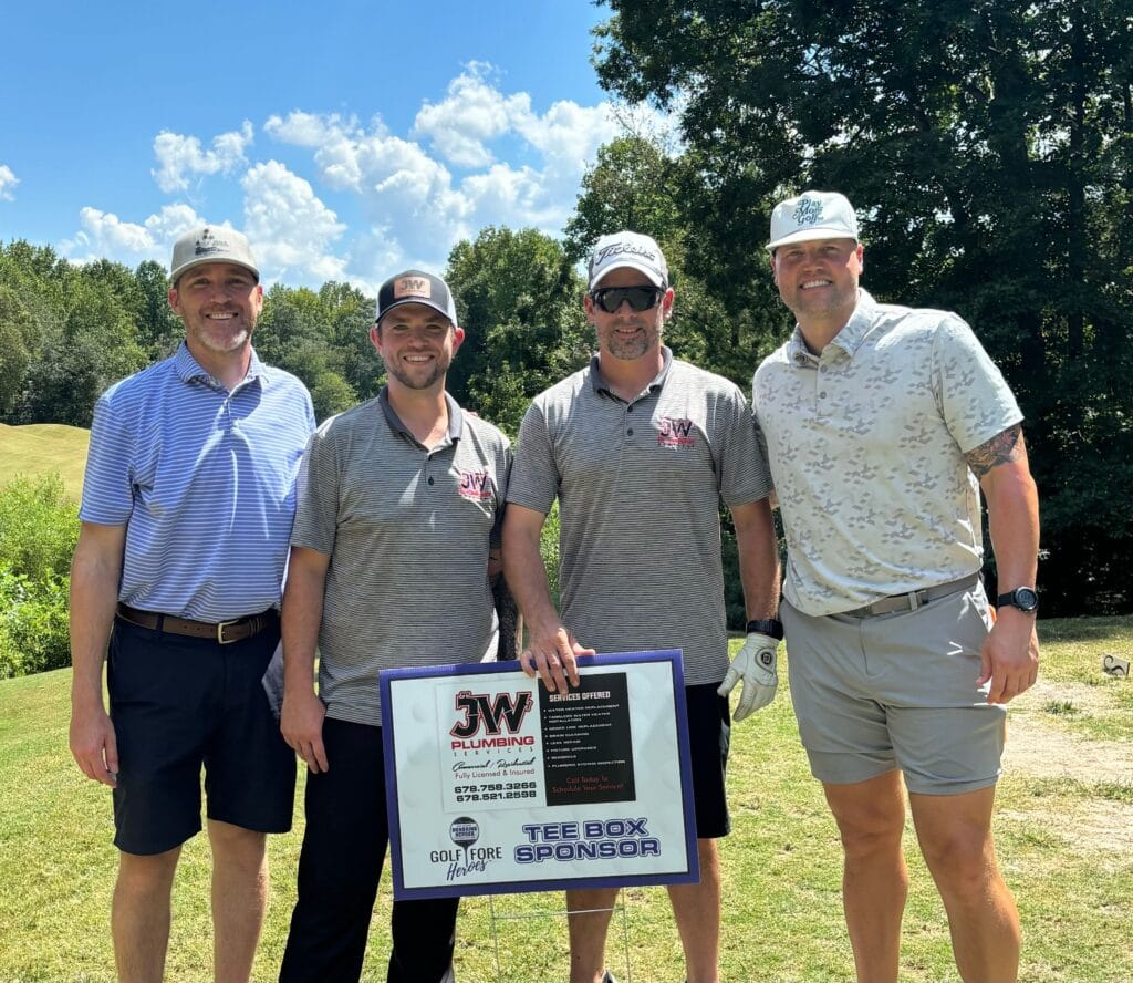 JW Plumbing Pros team members posing on a golf course, holding a sign promoting their plumbing services as a tee box sponsor.