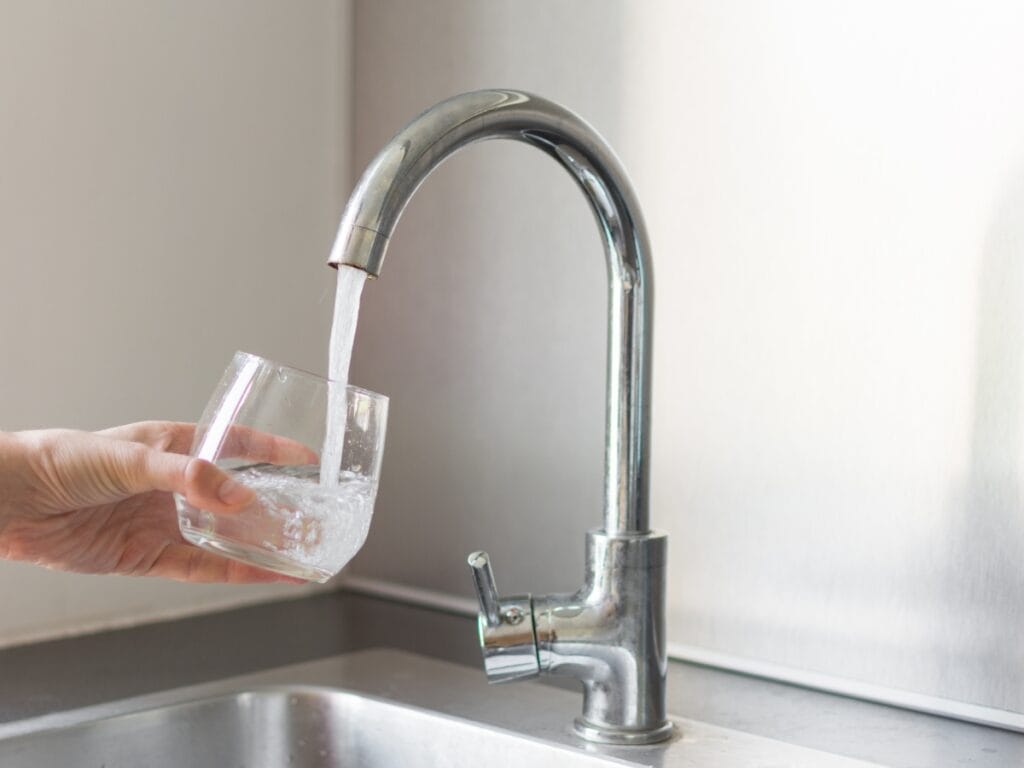 Hand holding a glass under a kitchen faucet with running water, illustrating water pressure and flow issues related to home plumbing.