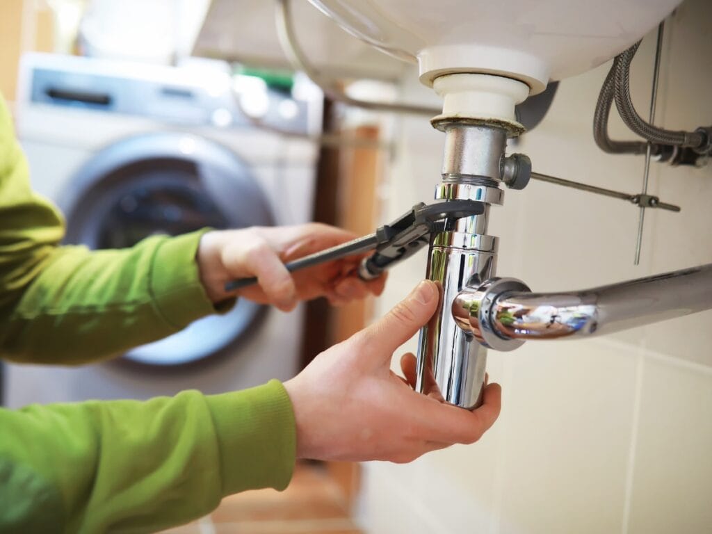 Person using adjustable wrench to tighten plumbing under sink, with washing machine in background, illustrating DIY plumbing maintenance for low water pressure issues.