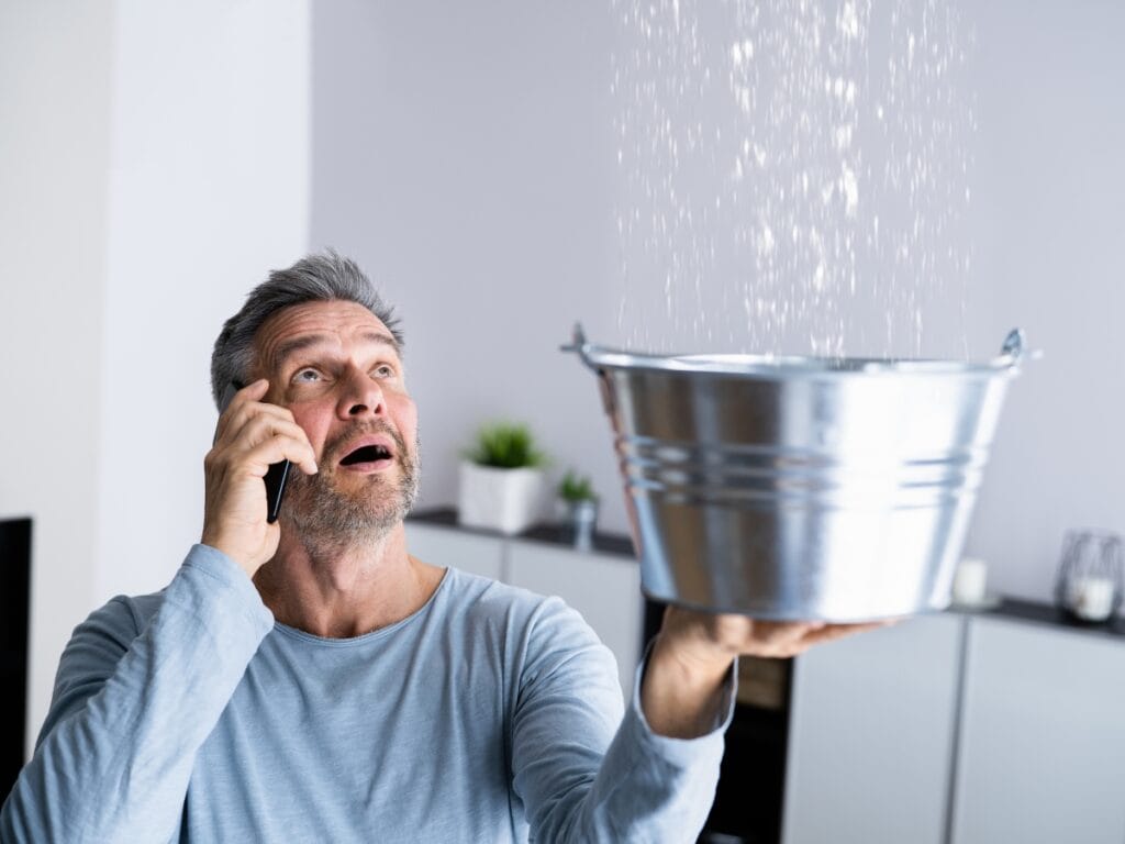 Man on phone looking up at water leaking into a metal bucket, illustrating signs of a hidden water leak in a home.