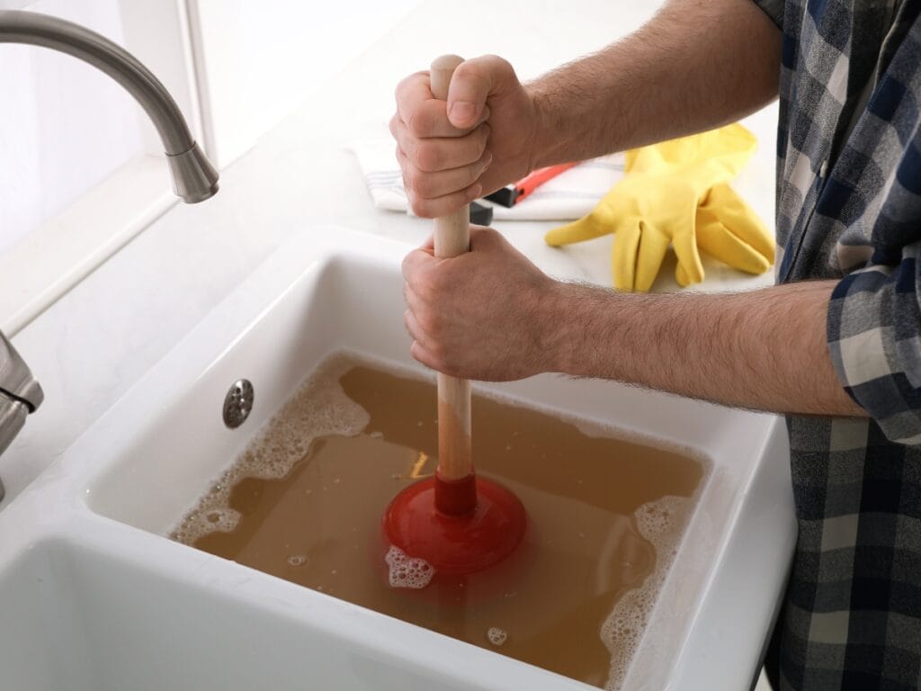 Person using a plunger to unclog a kitchen sink filled with dirty brown water; yellow rubber gloves rest nearby on the counter—a common scene when dealing with recurring drain clogs.