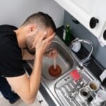 A man holding a plunger tries to unclog a kitchen sink, appearing frustrated by recurring drain clogs, surrounded by dirty dishes and cleaning items.