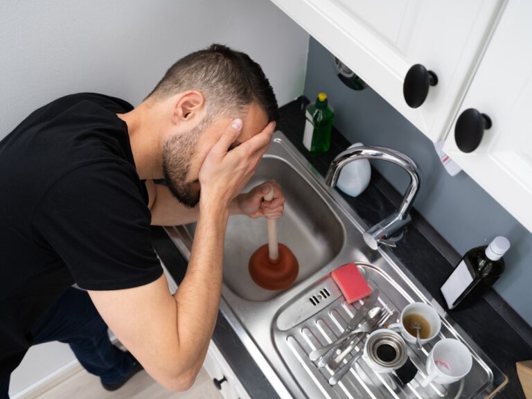 A man holding a plunger tries to unclog a kitchen sink, appearing frustrated by recurring drain clogs, surrounded by dirty dishes and cleaning items.
