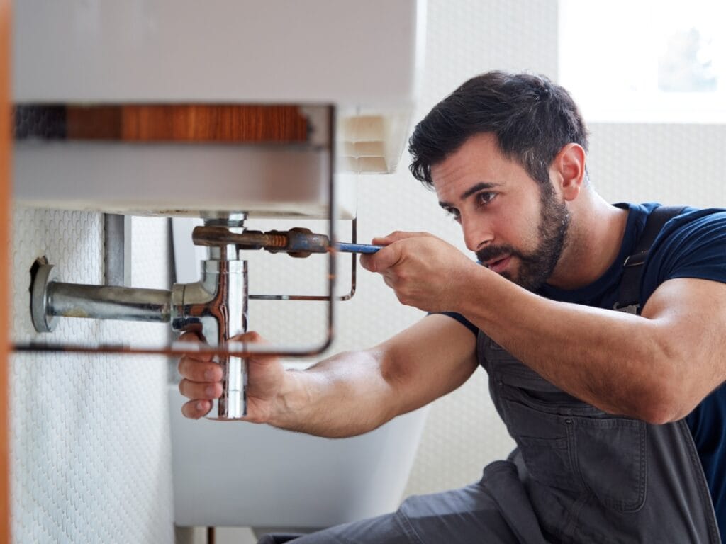 A plumber wearing overalls uses a wrench to tighten a pipe under a sink in a bathroom, working to fix recurring drain clogs and restore proper water flow.
