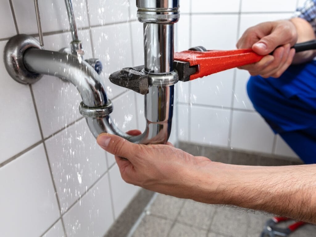 Person using a pipe wrench to fix a leaking pipe under a sink, with water spraying from the joint onto tiled wall and floor—a crucial repair to help stop drain clogs before they cause even more trouble.