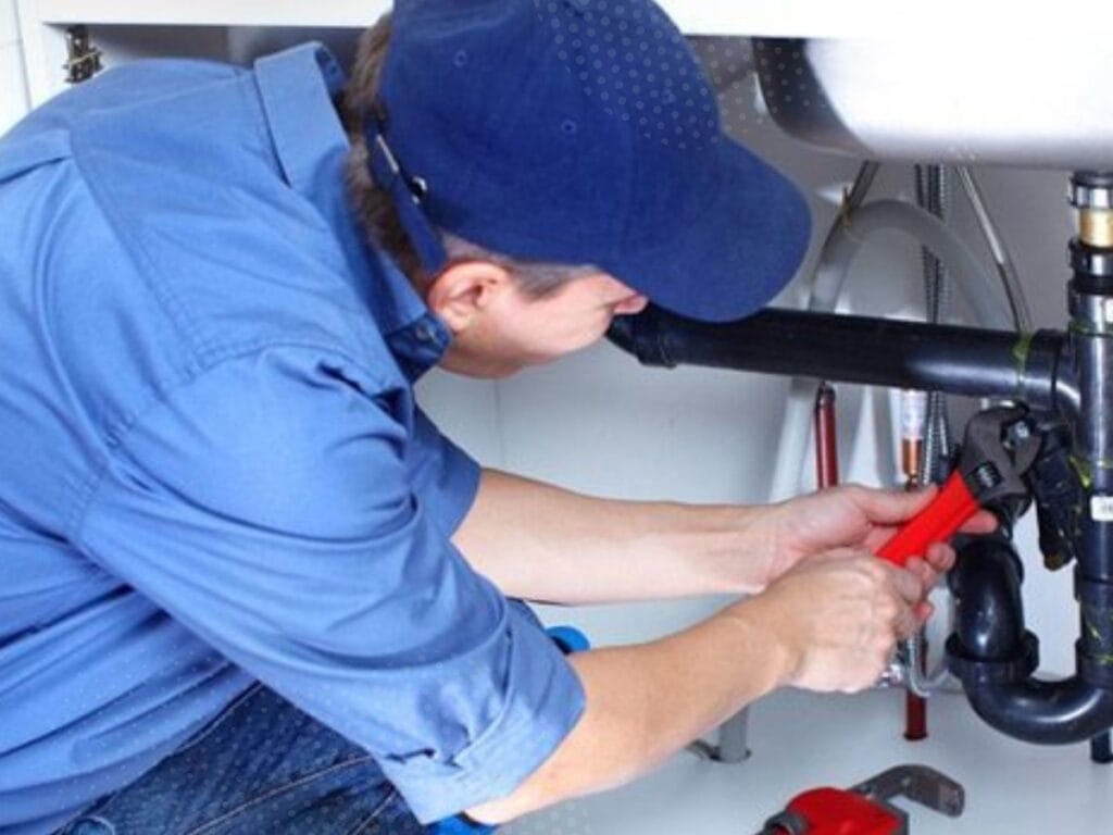 A person wearing a blue uniform and cap uses a red pipe wrench to repair plumbing under a sink, focusing on how to stop drain clogs and prevent recurring drain clogs.