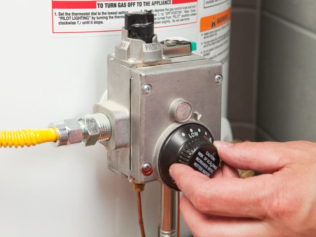 A hand adjusts the temperature dial on a water heater gas control valve, with warning labels visible—perfect for those deciding between water heater repair or replacement.