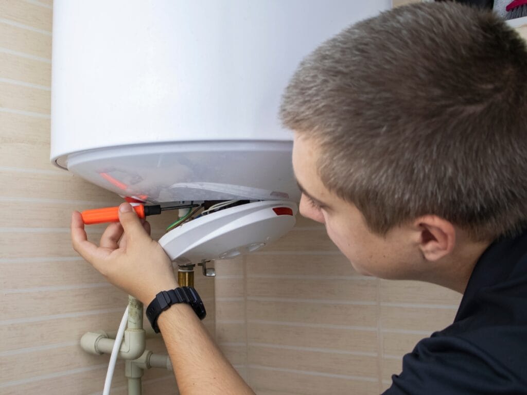 A person uses a screwdriver to inspect or perform water heater repair on the underside of a wall-mounted unit in a tiled room, helping to decide if water heater replacement is needed.