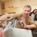 A man tries to solve plumbing problems as water sprays from a leaking kitchen faucet, while a concerned woman stands in the background talking on the phone.