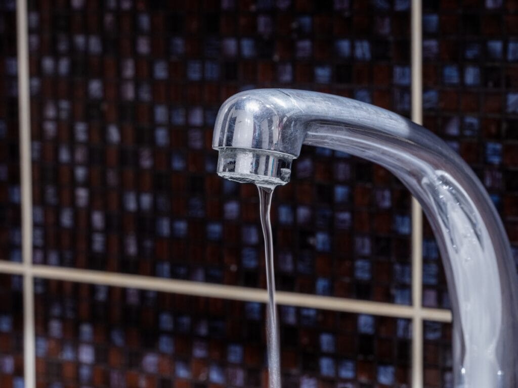 Common Plumbing Problems in Old Houses and How to Solve Them 9 A close-up of a chrome faucet with a thin stream of water flowing, set against a dark tiled background—an image often seen when dealing with plumbing problems in old houses.