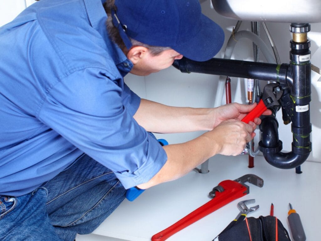 Common Plumbing Problems in Old Houses and How to Solve Them 10 A plumber in a blue uniform uses a pipe wrench to tighten a black pipe under a sink, offering expert plumbing solutions for old houses, with various tools laid out nearby.