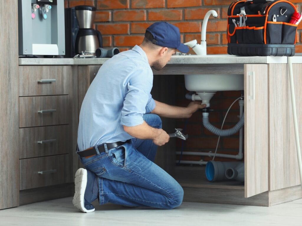 Common Plumbing Problems in Old Houses and How to Solve Them 11 A plumber kneels to solve plumbing problems under a kitchen sink, holding a wrench, with tools in a bag on the counter and a brick backsplash in the background—perfect for tackling issues common in old houses.