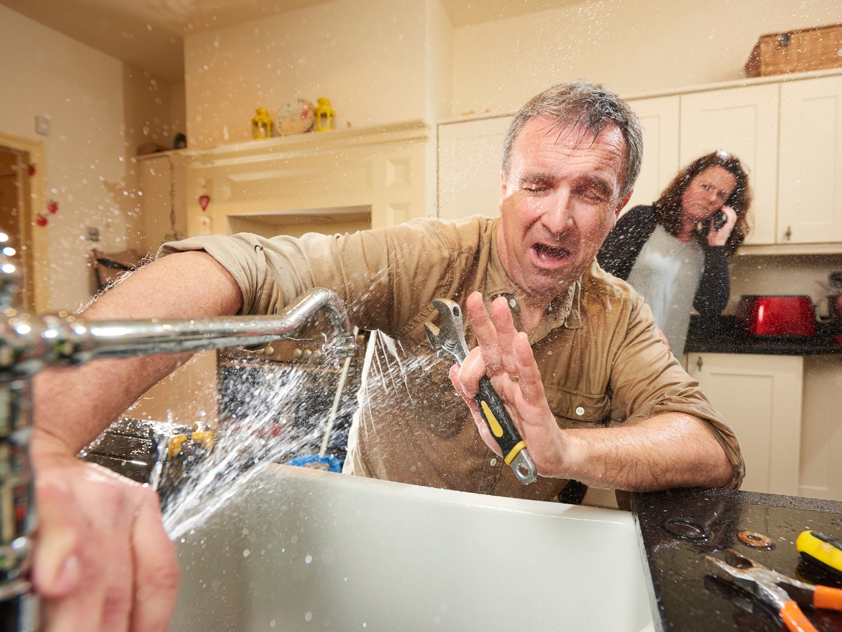 A man tries to solve plumbing problems as water sprays from a leaking kitchen faucet, while a concerned woman stands in the background talking on the phone.
