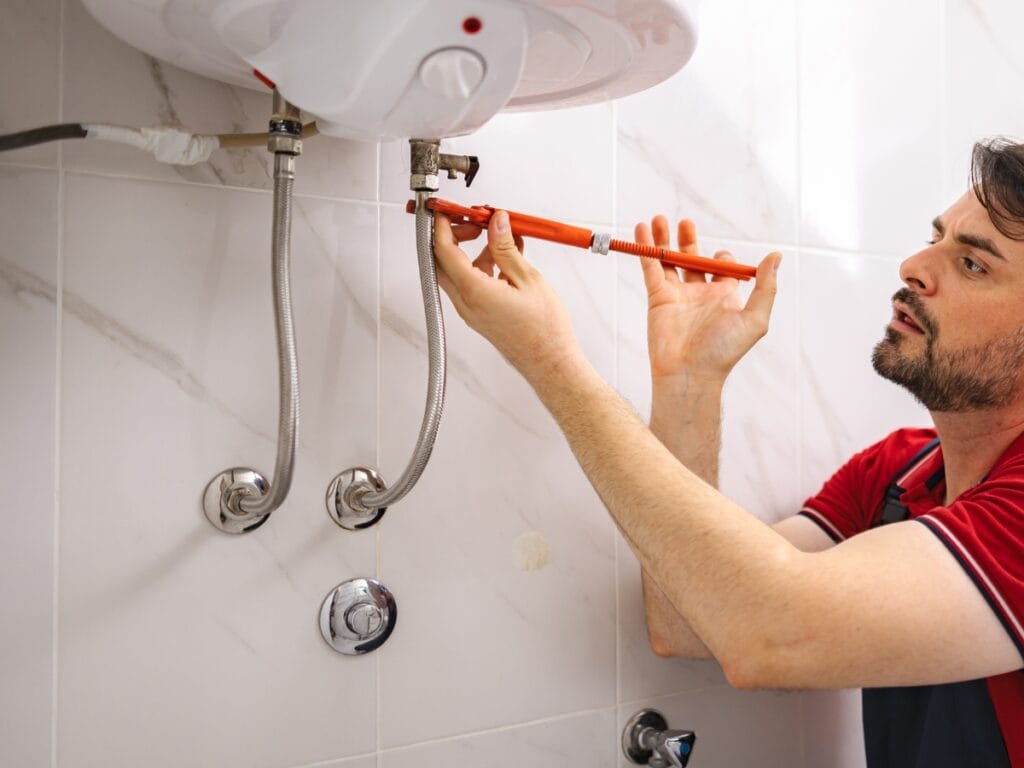 Benefits of Tankless Water Heaters for Energy-Efficient Homes 6 A man uses a wrench to tighten or loosen a valve on a wall-mounted tankless water heater in a tiled bathroom, ideal for energy-efficient homes.