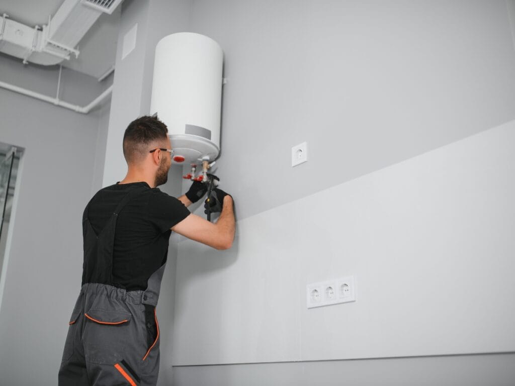 Benefits of Tankless Water Heaters for Energy-Efficient Homes 11 A man in work overalls installs or repairs a white tankless water heater mounted on a gray wall in a modern room, enhancing comfort in energy-efficient homes.